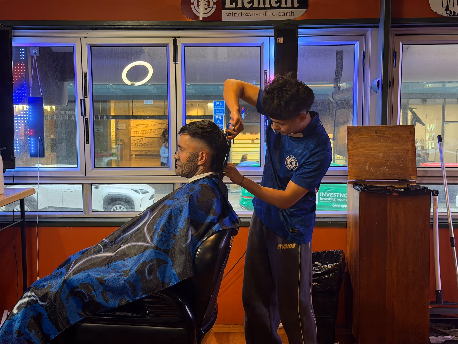 Barber cutting a customer’s hair inside a shop with large windows showing cars outside.