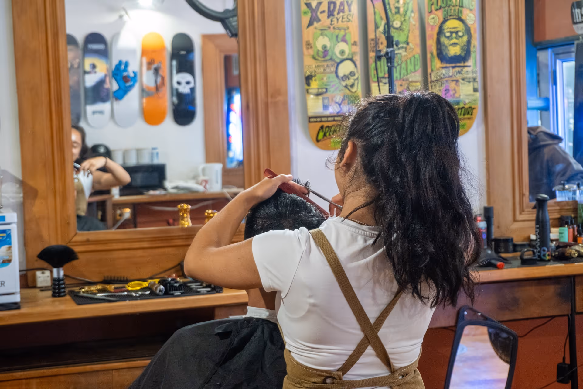 Female barber with ponytail cutting a male client's hair in a studio with skateboard decks on the wall.