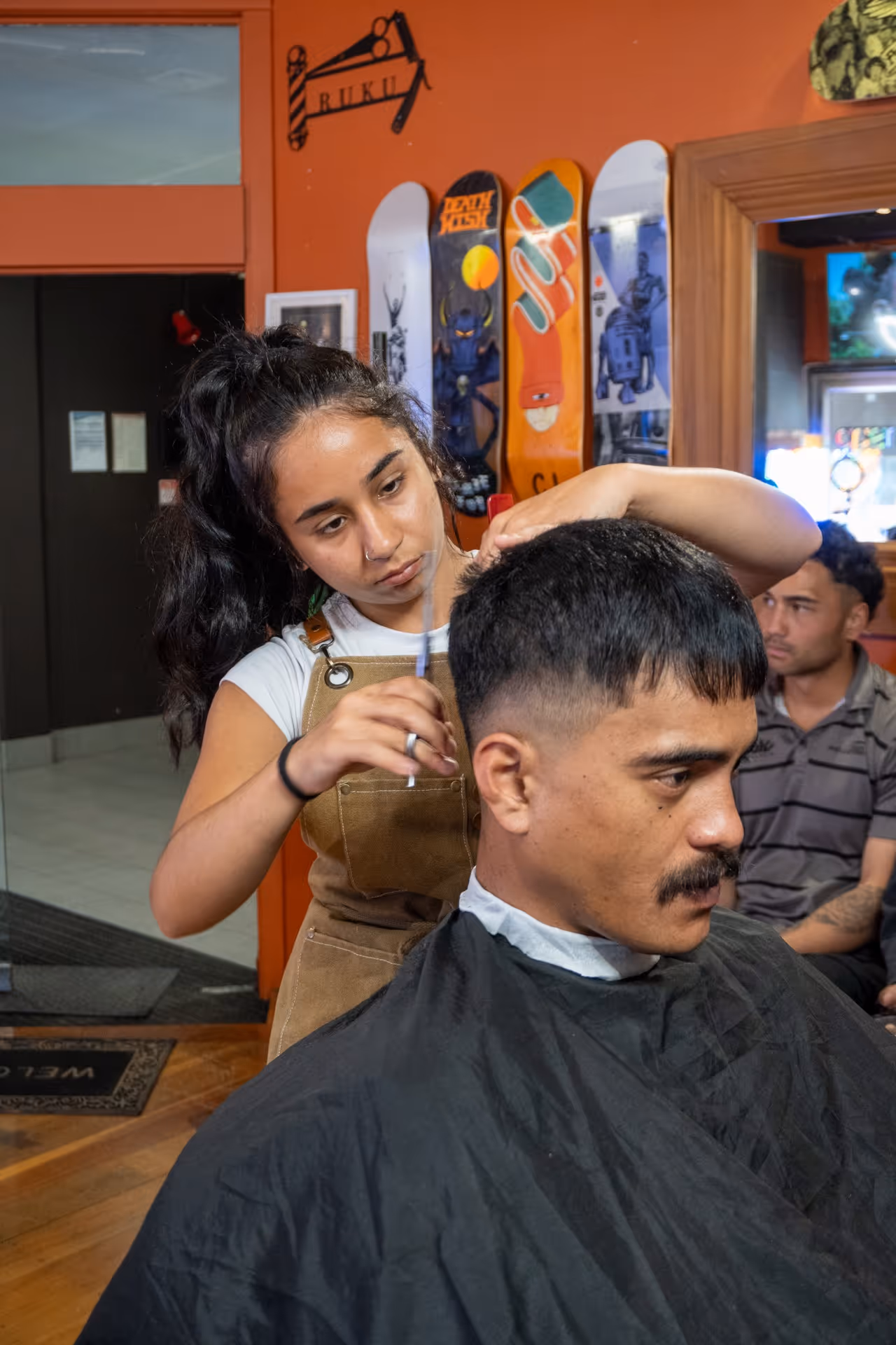 Female barber in brown apron cutting a man's short black hair inside a barbershop with orange walls and skateboards mounted on the wall.