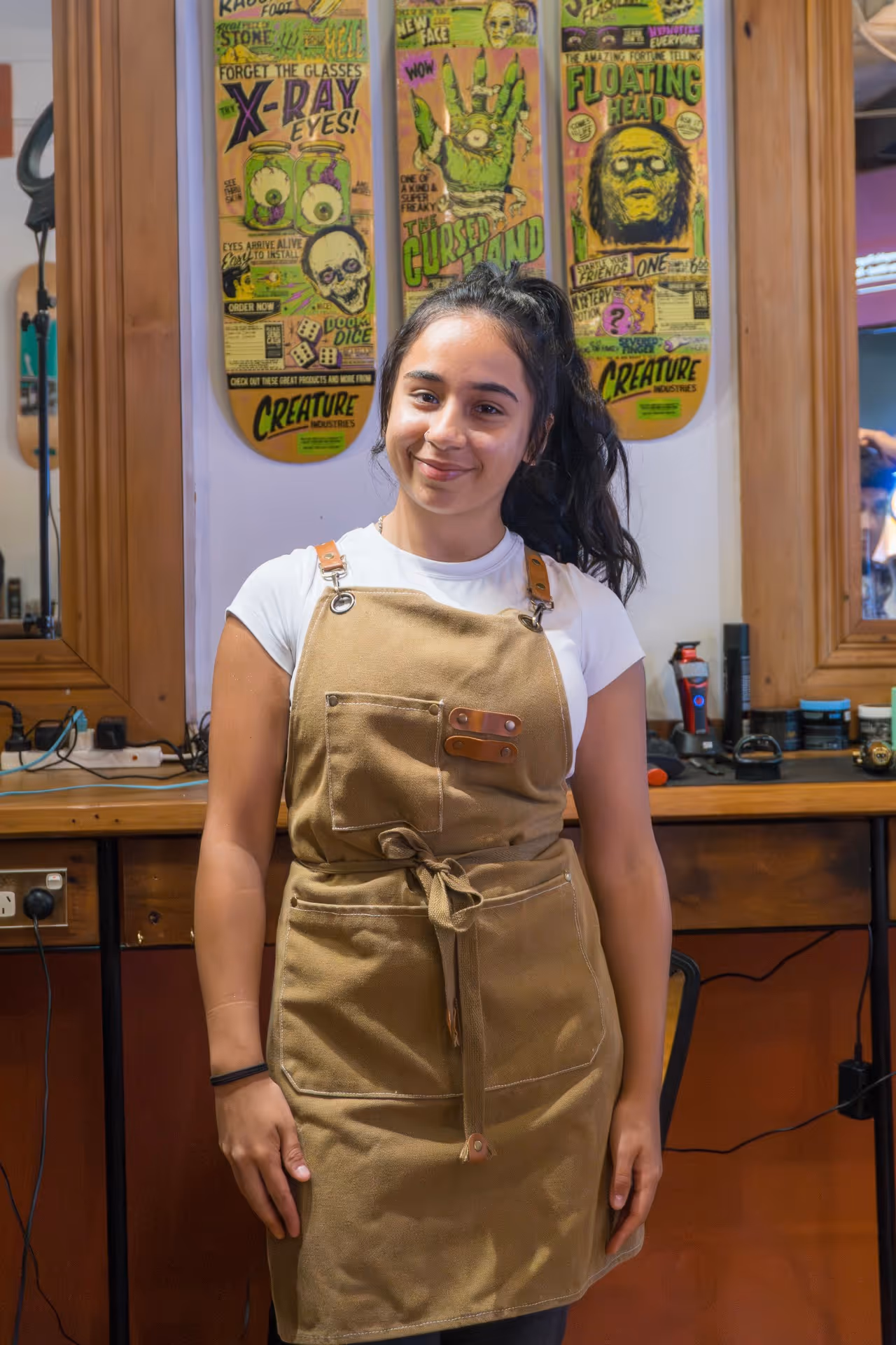 Smiling woman with dark hair in a ponytail wearing a tan apron over a white shirt, standing in a barbershop with wooden mirrors and colorful posters behind her.